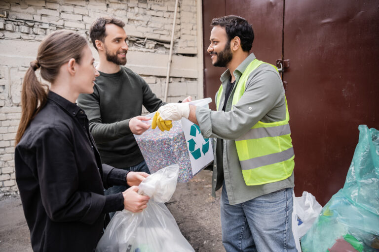 Smiling volunteer giving trash bin with recycle sign to indian worker in safety vest and gloves